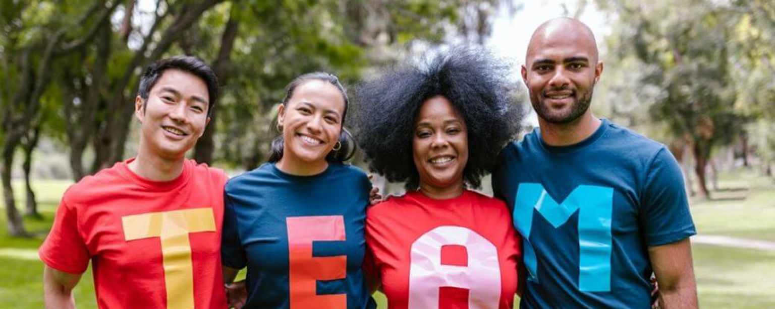 Vier lachende Mitarbeitende tragen T-Shirts mit Buchstaben, die das Wort „TEAM” ergeben 